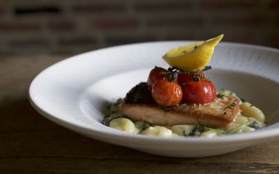 Salmon fillet on a bed of creamy new potatoes with roasted vine cherry tomatoes and lemon. Served in a white dish and photographed against a red brick wall.