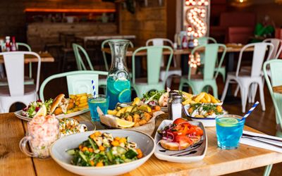 wooden brown table laid out with different dishes and blue lagoon cocktails at the Ohso Brighton. Featured in our places to eat in Brighton