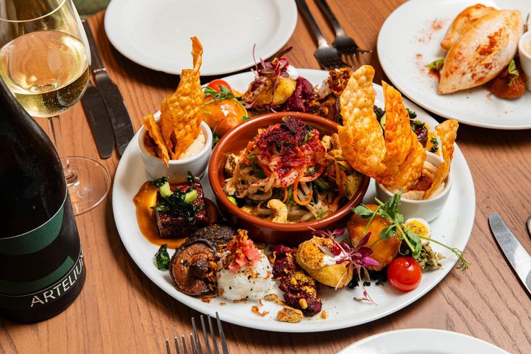 A vibrant and beautifully arranged sharing platter featuring an assortment of colourful and artistically plated small bites, including crispy wonton chips, fresh vegetable salad, glazed mushrooms, sushi rice, and gourmet appetizers. The dish is complemented by a bottle of sparkling wine and a glass of white wine, set on a wooden table with cutlery and additional plates in the background, creating a visually inviting dining experience.