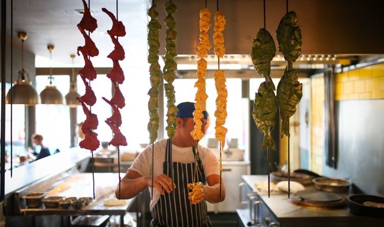 The tandoor oven area. Pictured an Indian chef preparing skewers of fish, chicken and lamb. The skewers have bright marinade sauces which are red, yellow, green. 