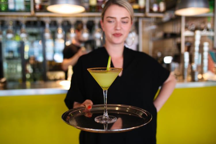 Beautiful young lady and waitress holding a single martini cocktail on a silver tray. A yellow out of focus wall in the background. She has nail varnish on her hands and she is wearing a black dress. 