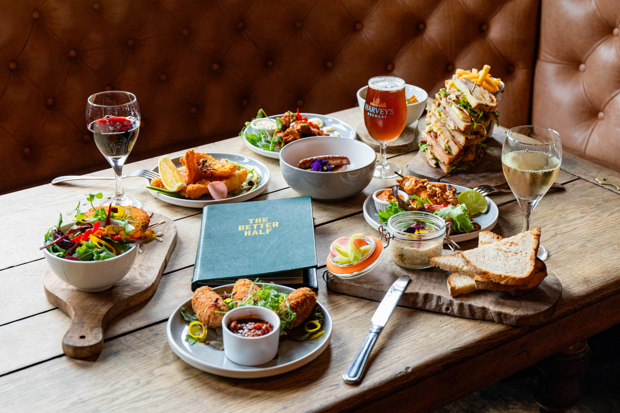 diner table laid out with seven different dishes including big sandwich, meat, beer and wine