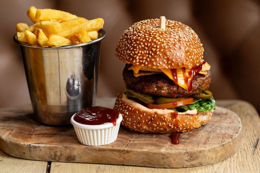 delicious looking meat burger with sauces dripping on the side served with chips on the side, all on the brown wooden board.