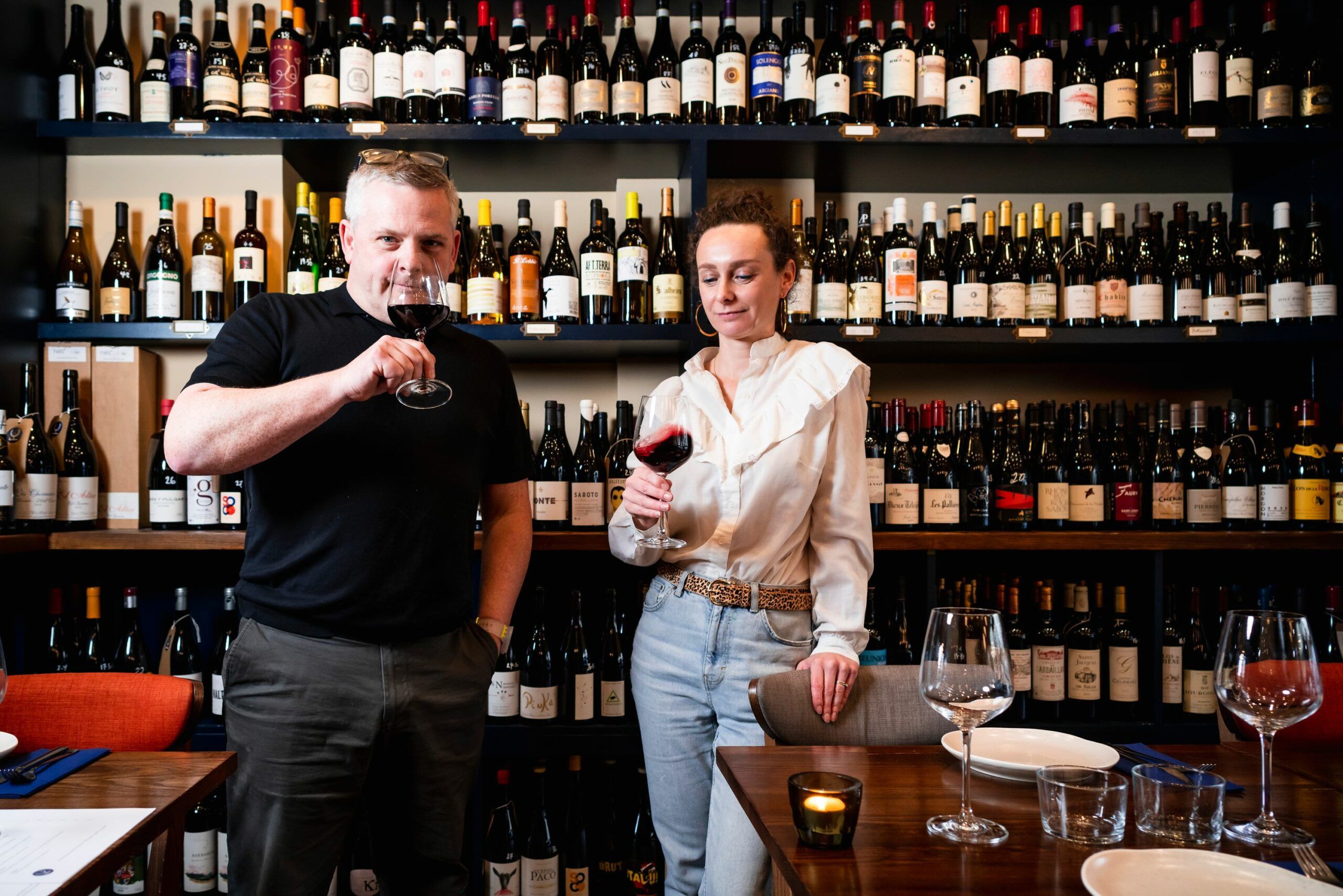 team having glass of wine against a back drop of wine bottles. Fourth and Church, wine shop and deli in Hove