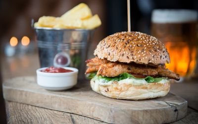 A chicken burger and chips with a pot of sauce served on a wooden board alongside a pint of beer.