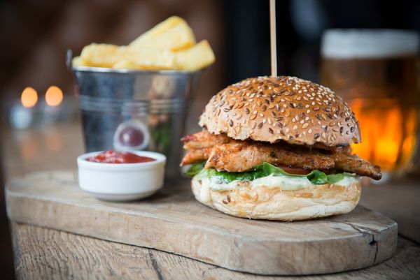 A chicken burger and chips with a pot of sauce served on a wooden board alongside a pint of beer. Pub dinner
