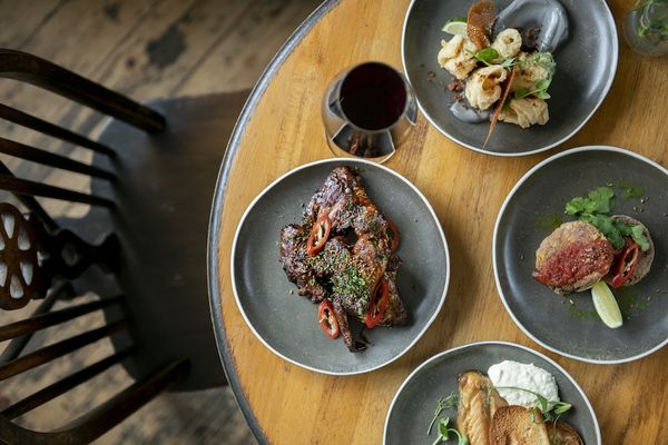 A selection of meals photographed overhead on a wooden table with a black wooden chair. All served on grey ceramic plates with a glass of red wine.