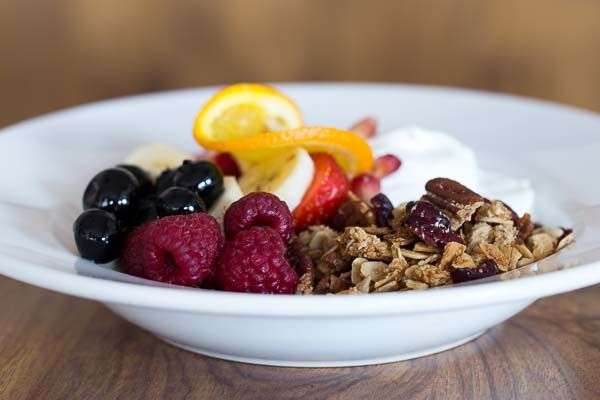 Breakfast bowl of granola, fresh raspberries and blueberries served alongside yoghurt in shallow white bowl.