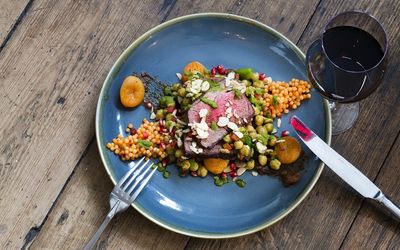 A blue ceramic plate with a bed of giant couscous, sliced rare meat, apricots and glass of red wine.