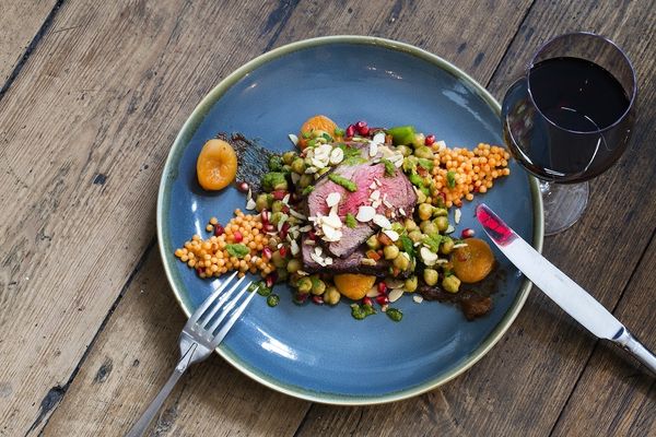 A blue ceramic plate with a bed of giant couscous, sliced rare meat, apricots and glass of red wine.