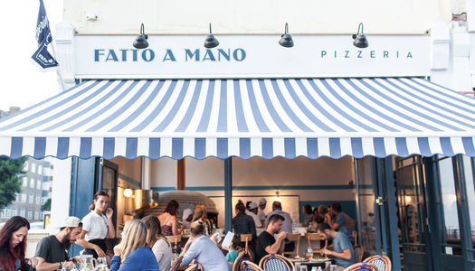 Diners enjoying pizza in the alfresco area of Fatto A Mano in Hove. With stripy white and blue roll our sun shade against the backdrop of a white building in Hove. Fatto a Mano Hove • Neapolitan Pizza in the Heart of Hove