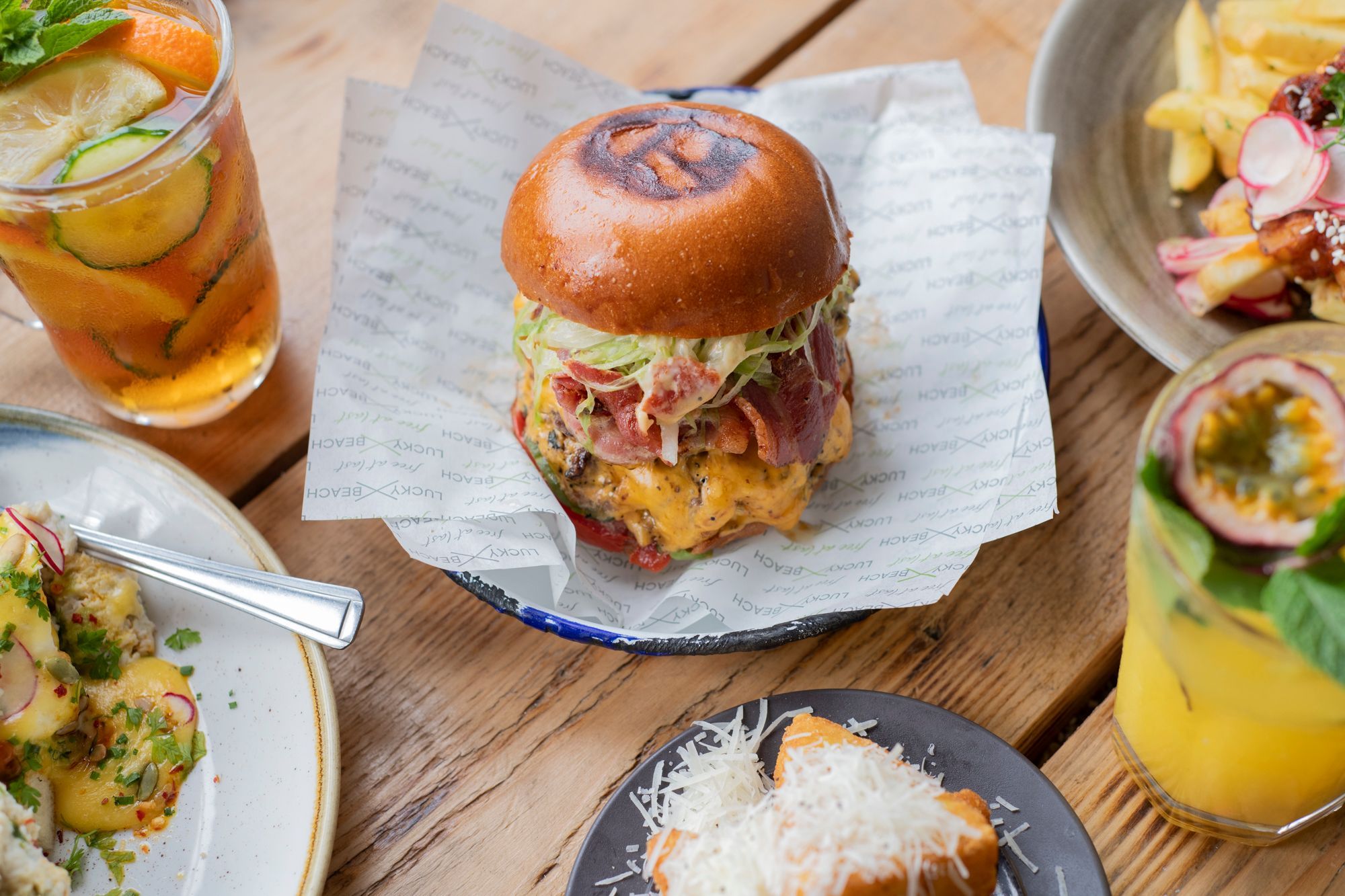 Lucky Beach Brighton burger sat among several dishes which you cant quite see. The burger has lots of bacon and cheese relish. There is a summer drink to the side, 