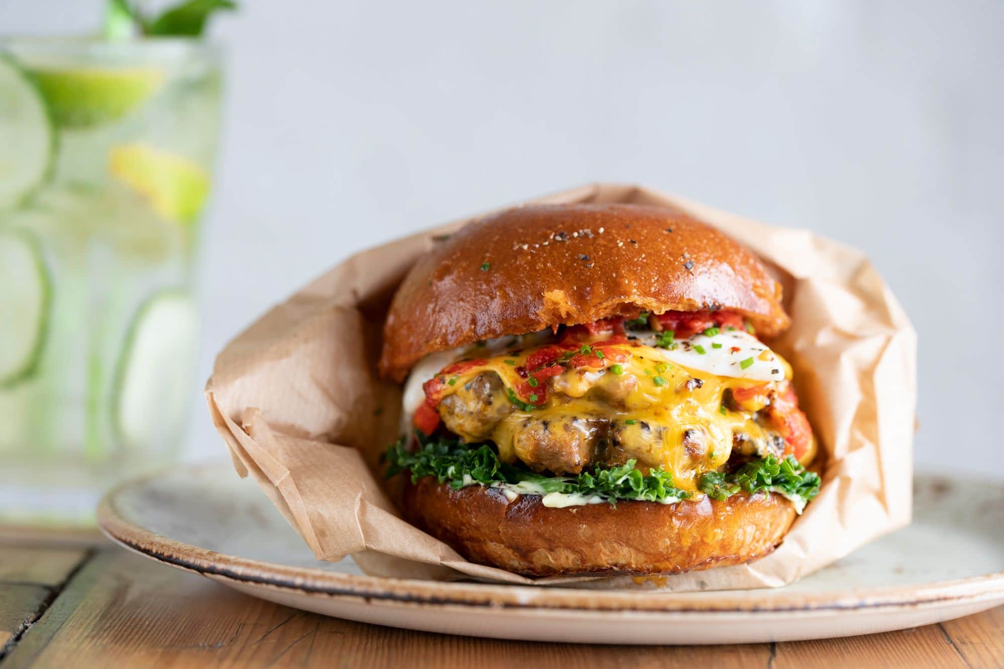 Lucky Beach Brighton. Burger sat in brown paper, with cheese oozing out. A double pattie with relish, on a speckled clay plate with a white wall backdrop and a jug of water