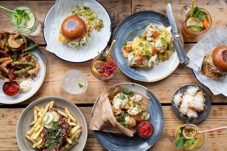 A Brighton breakfast of dazzling proportions. An overhead shot of several breakfast dishes placed on a wooden table. Included are a breakfast burger, scrambled eggs, granola bowls, cocktail drinks and more. a great breakfast option by the Brighton seafront. Part of the Brighton seafront restaurants guide.