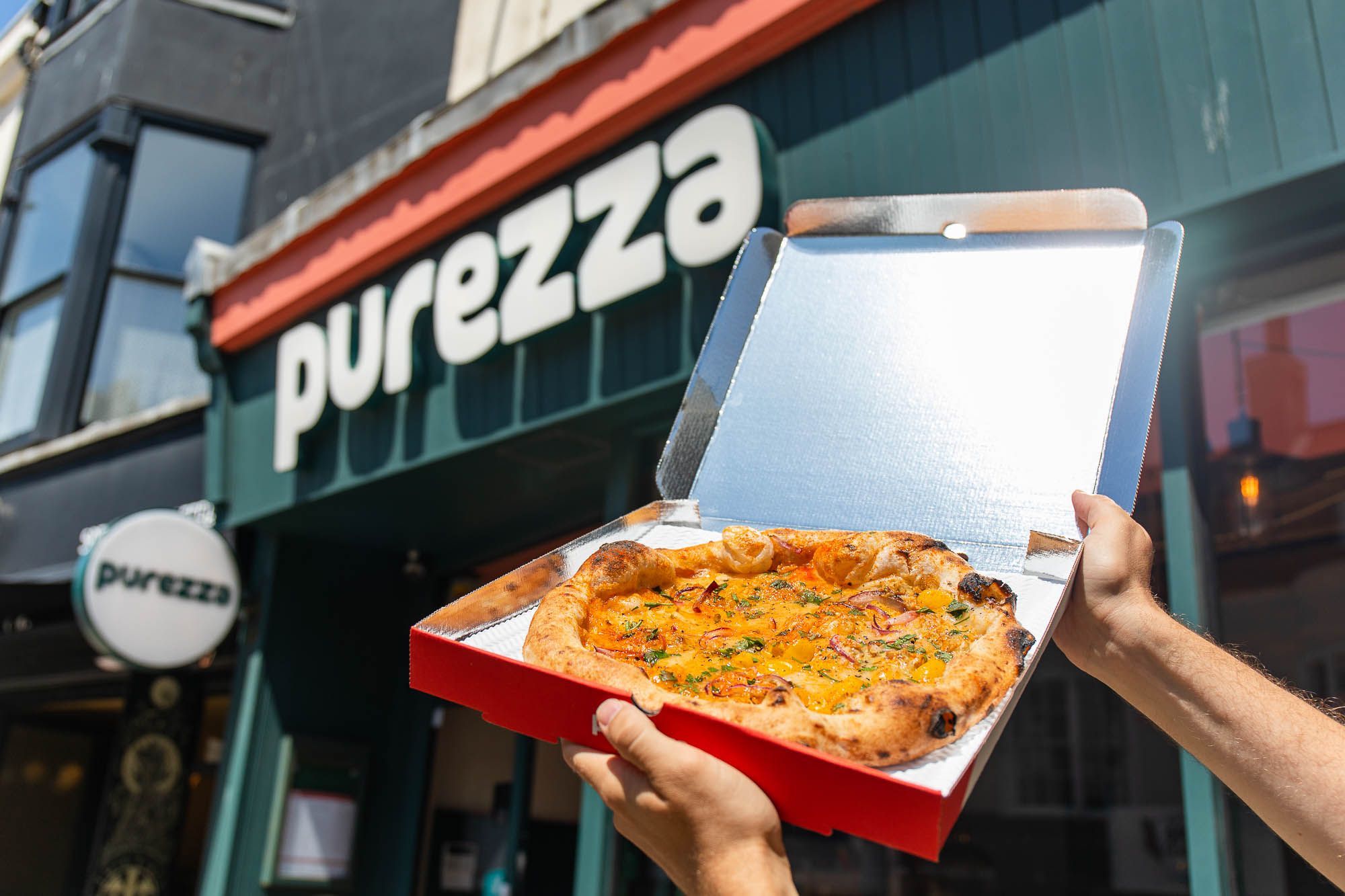 hands holding open box of purezza pizza next to entrance sign Purezza