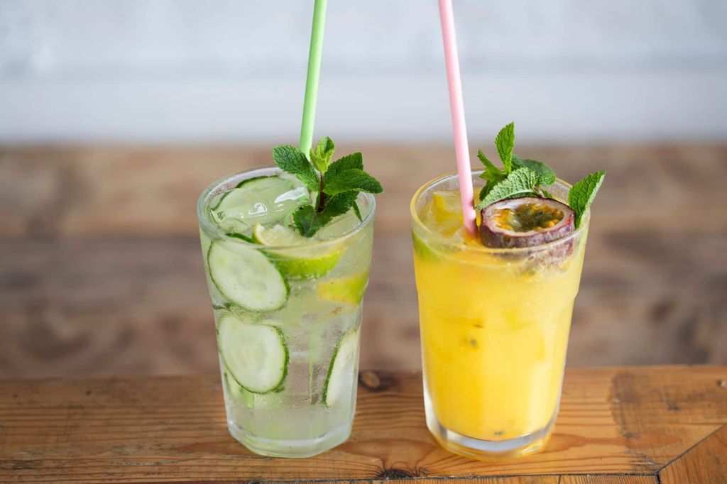Alcohol free cocktails in Brighton. one orange based drink, one lime based. Being carried on a tray with a beach and blue sky back ground. The location is Brighton beach on Brighton seafront.