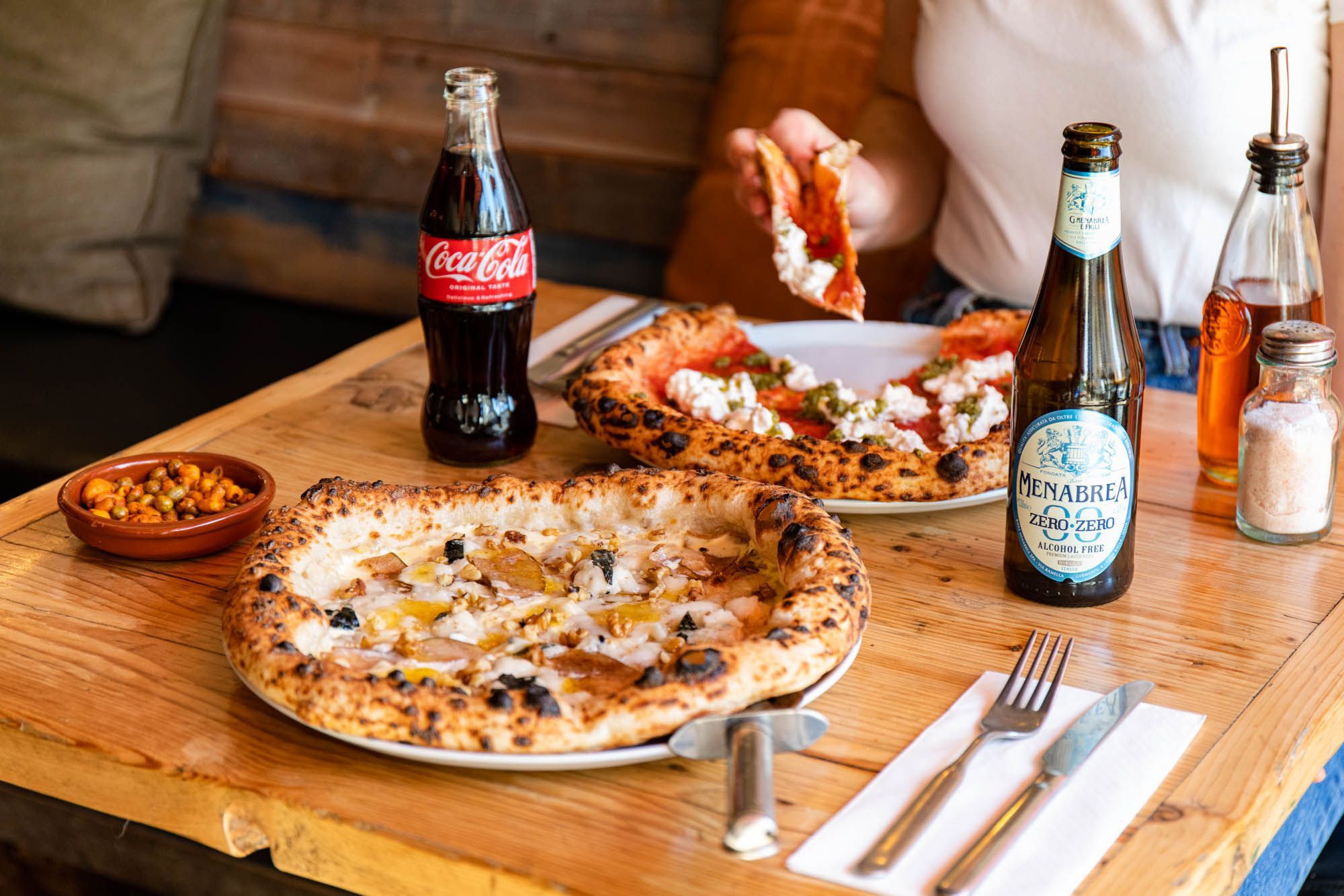 table laid out with two pizzas, one of the pizzas is being eaten by person, for the drinks they served beer and Coca Cola