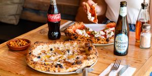 table laid out with two pizzas, one of the pizzas is being eaten by person, for the drinks they served beer and Coca Cola