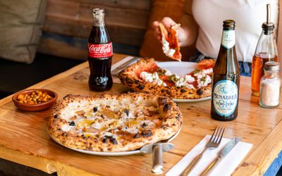 table laid out with two pizzas, one of the pizzas is being eaten by person, for the drinks they served beer and Coca Cola