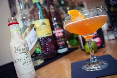 An orange cocktail photographed on the bar with bottles of alcohol in the background.