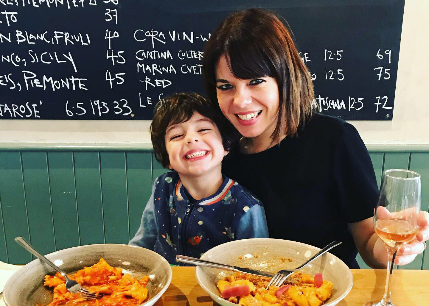 A lady and child sat at at table eating pasta with a chalkboard menu behind them.