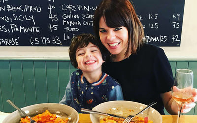 A lady and child sat at at table eating pasta with a chalkboard menu behind them.