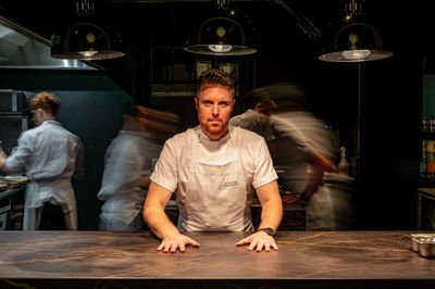 etch Brighton. Steven Edwards standing at the pass at his Hove flagship restaurant which is called etch. Steven is wearing a white chefs apron and other chefs are working behind him and they are slightly faded out into a darkness. Steven Edwards has 3 AA rosettes, he is a MasterChef winner and he is featured in the Michelin guide. Fine Dining Brighton. Steven Edwards will appear at the 2024 Brighton Foodies Festival.