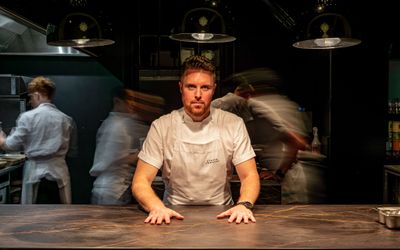 etch Brighton. Steven Edwards standing at the pass at his Hove flagship restaurant which is called etch. Steven is wearing a white chefs apron and other chefs are working behind him and they are slightly faded out into a darkness. Steven Edwards has 3 AA rosettes, he is a MasterChef winner and he is featured in the Michelin guide. Fine Dining Brighton. Steven Edwards will appear at the 2024 Brighton Foodies Festival.