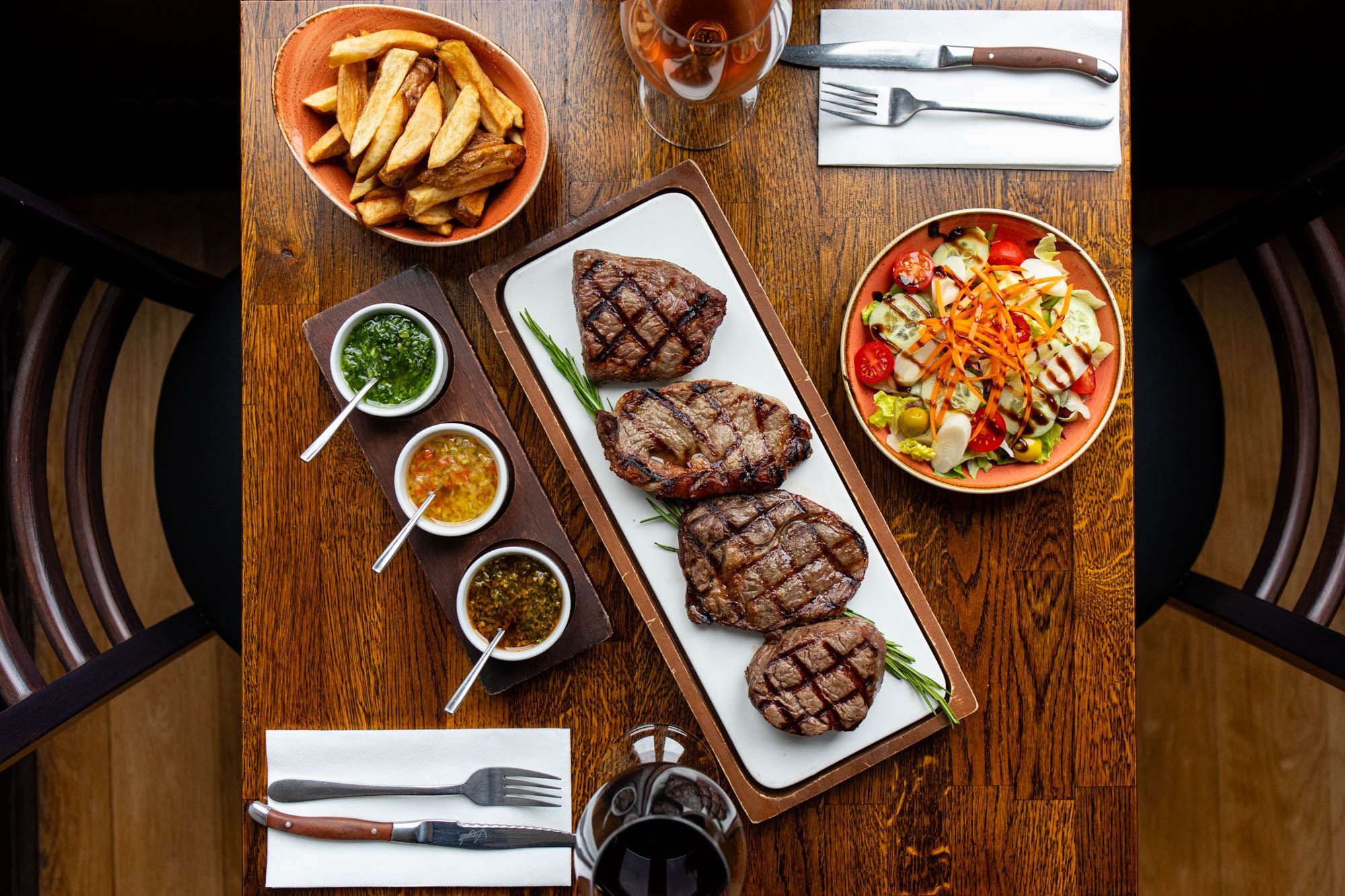 overhead shot of the brown wooden table at LatinoAmerica Hove, laid out with plate of Argentinian steak, different sauces, chips and salad