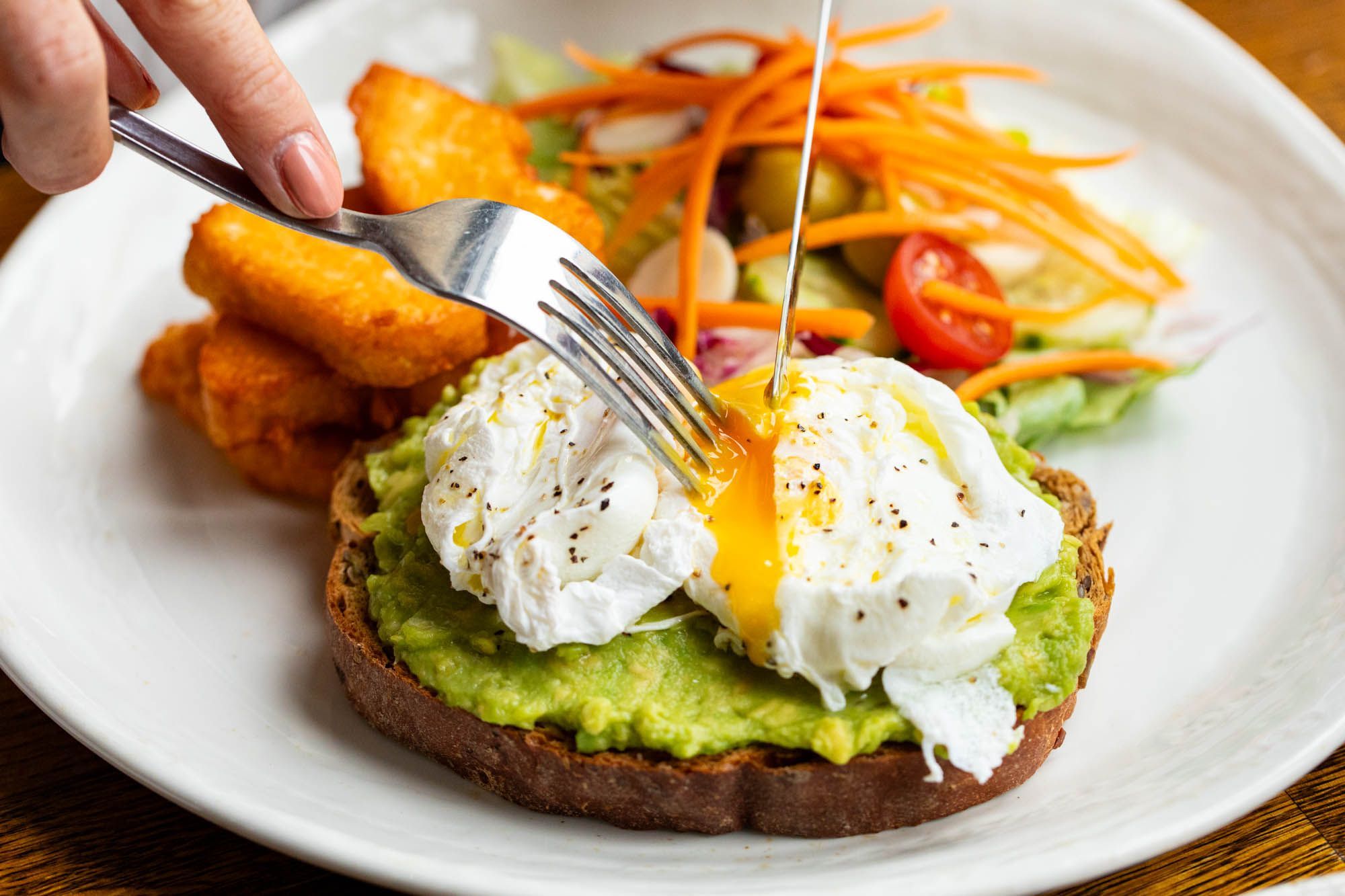 close up shot of knife and fork cutting trough egg on the avocado toast served with nuggets on the white plate