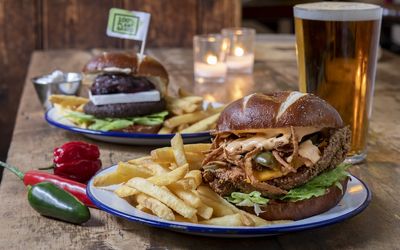 Enamel plates of burgers and fries served on a wooden dining table with tealights and a pint of beer.