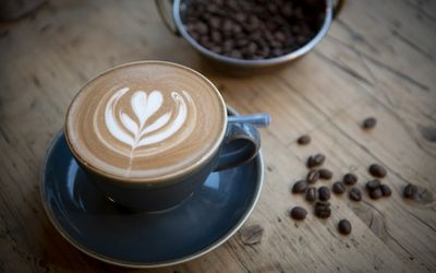 Trading Post Coffee. Available for Brighton takeaway and home delivery. Pictured, a flat white on a wooden table with roasted coffee beans in the background.