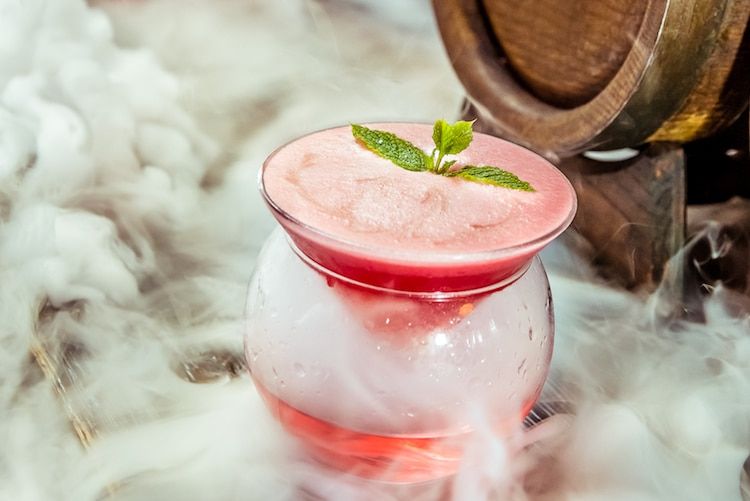 A pink cocktail with a herb garnish sat inside another glass with liquid nitrogen steam.