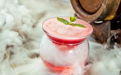 A pink cocktail with a herb garnish sat inside another glass with liquid nitrogen steam.