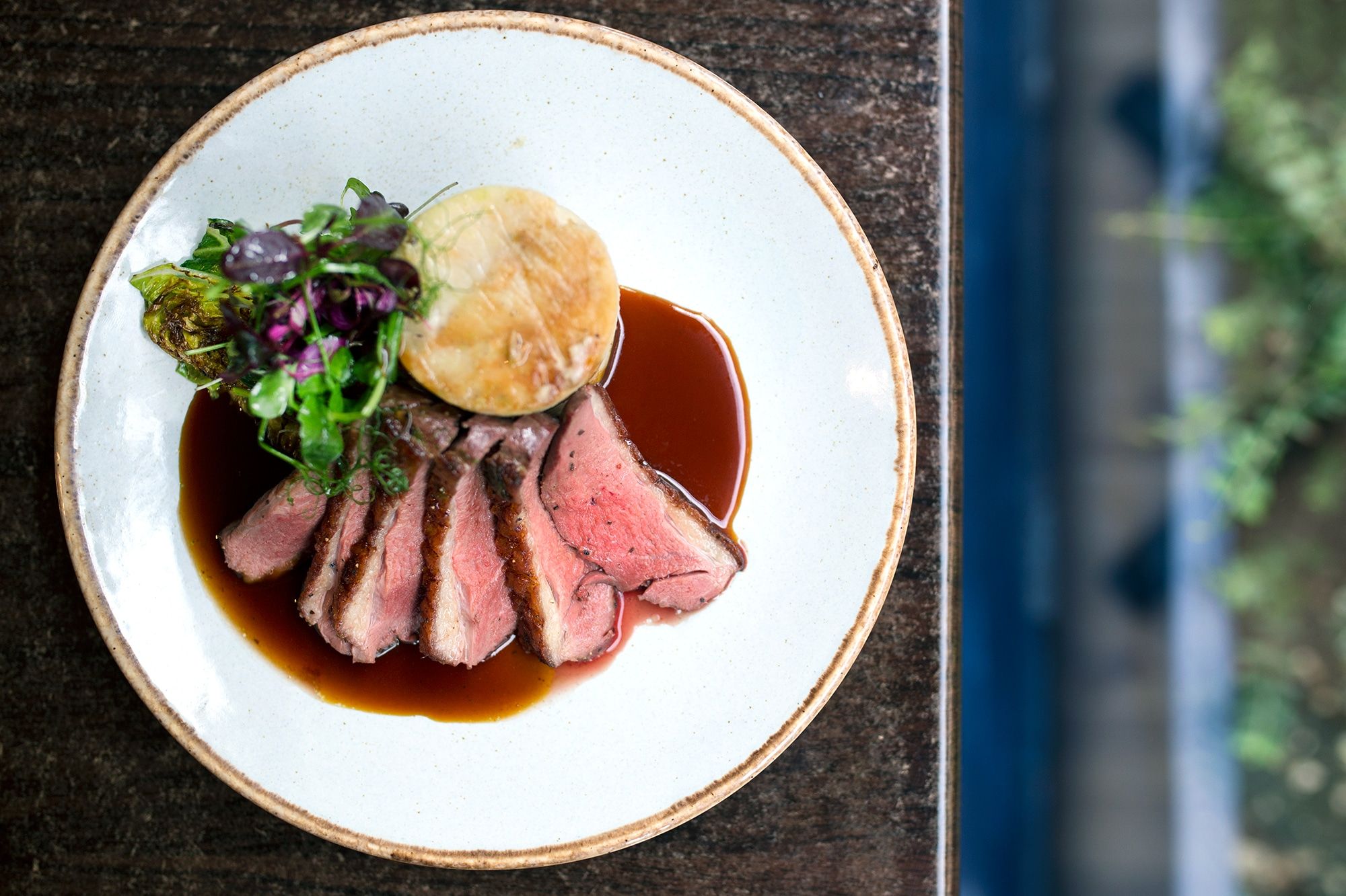 Sliced rare meat with fondant potatoes and a rich gravy. Served on a white ceramic plate with a brown glazed edge. Leisurely lunch ideas in Brighton