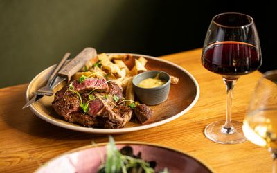 Steak frites with red wine A hearty plate of sliced medium-rare steak served with chunky chips and a pot of béarnaise sauce, accompanied by a glass of red wine on a wooden table.
