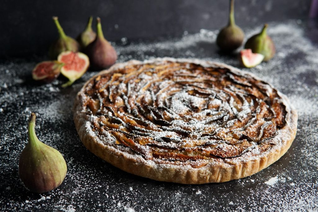 A caramel and fig tart on a grey surface dusted with icing sugar and figs arranged about it.