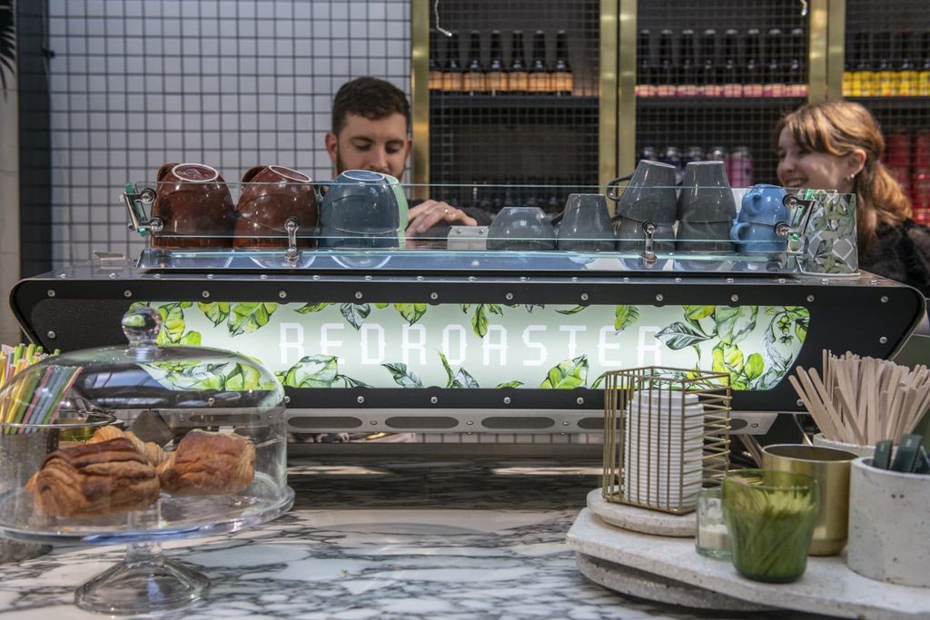 Redroaster cafe in Brighton. Pictured, two baristas behind their coffee machine, preparing coffee. A cake stand is in the foreground. The two staff members are smiling