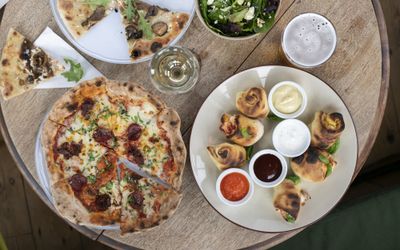 Overhead view of pizza, dough balls and sides on a round wooden table.