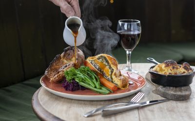 Gravy being poured over a Sunday roast, with roast wellington, Yorkshire pudding carrots, broccoli and cabbage. Red wine and a side of cauliflower cheese are in shot.