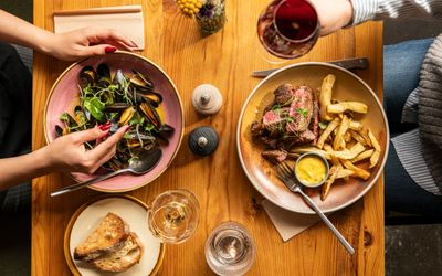 Shared dinner with steak and mussels An overhead shot of a shared meal including a bowl of mussels with herbs, a steak and fries plate with dipping sauce, sourdough bread, and glasses of wine, capturing a cozy dining moment.