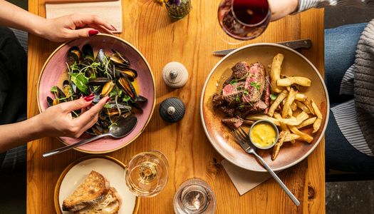 Shared dinner with steak and mussels An overhead shot of a shared meal including a bowl of mussels with herbs, a steak and fries plate with dipping sauce, sourdough bread, and glasses of wine, capturing a cozy dining moment.