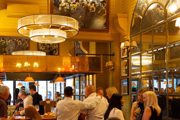 People gathered in a high ceilinged bar and restaurant with large mirrors and chandeliers, the central bar is polished metal and the words WINES, BEERS and COCKTAILS are written on them