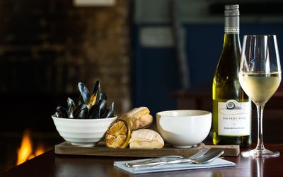 A platter of moules marinière, rustic bread and white wine. Displayed in front of an open fire.