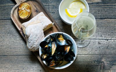 Mussels served in a bowl on a wooden board with fresh bread and grilled lemon. Photographed against a wooden table.