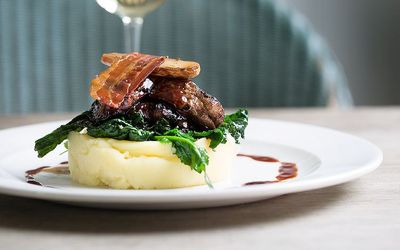 A disc of mashed potato topped with greens and crispy bacon. Photographed alongside a glass of wine.