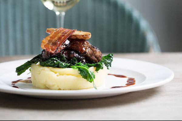 A disc of mashed potato topped with greens and crispy bacon. Photographed alongside a glass of wine.