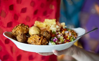 A large white dish being held up against a colourful fabric backdrop with curry, bhajis and salad.