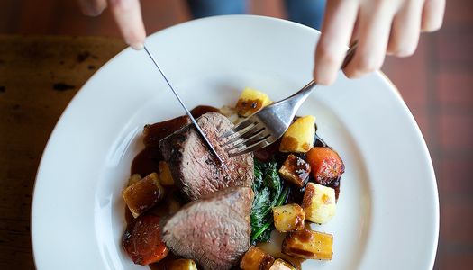 Sunday roast beef with cubed roasted root vegetables and a rich gravy. Hands featured in the shot holding a knife and fork.