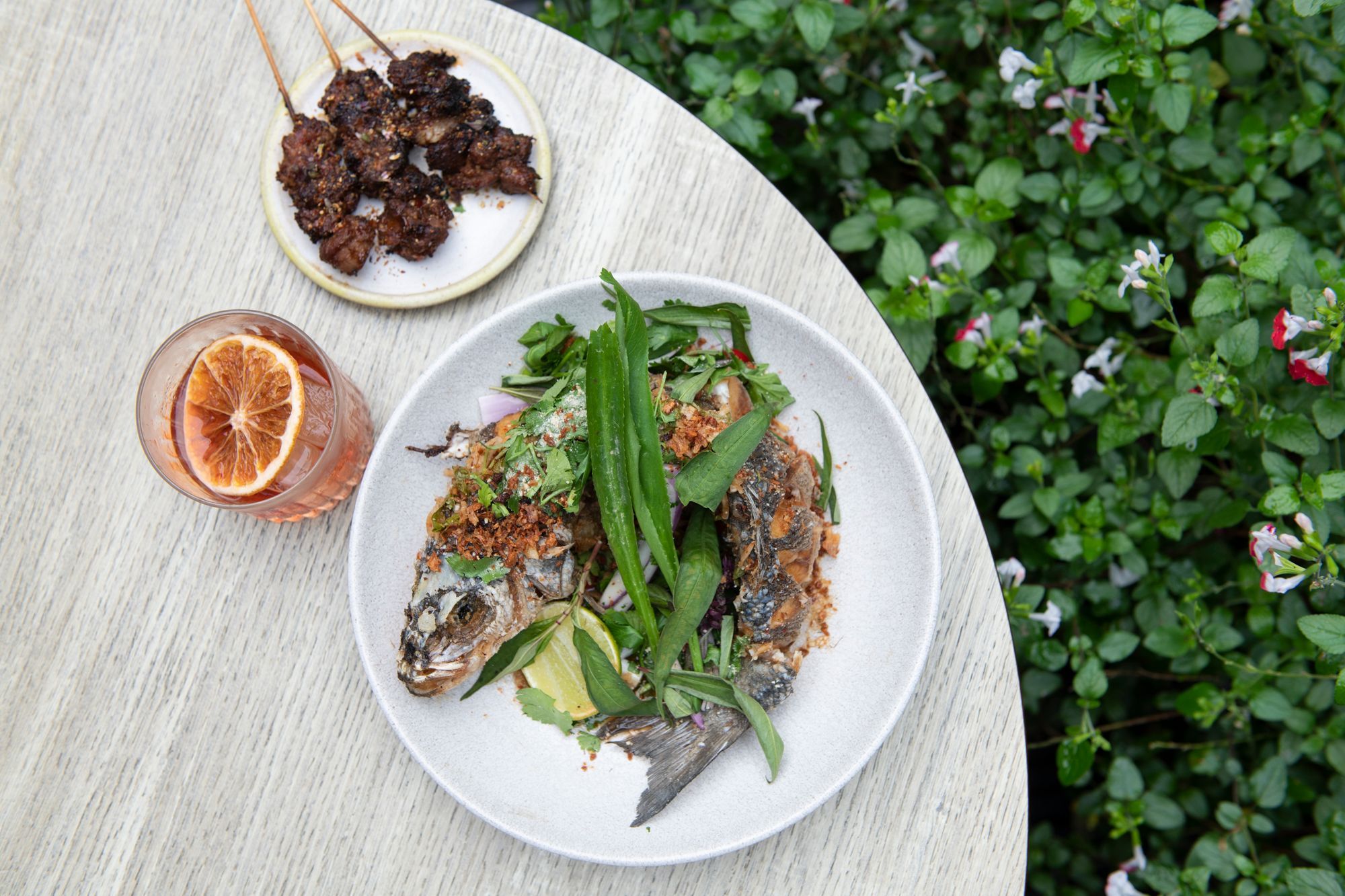 sea bass with glass of cocktail. A picture taken in the Lucky Khao Brighton rear terrace garden. The picture shows a grey table top with green garden plant to the right. It looks very pretty. 